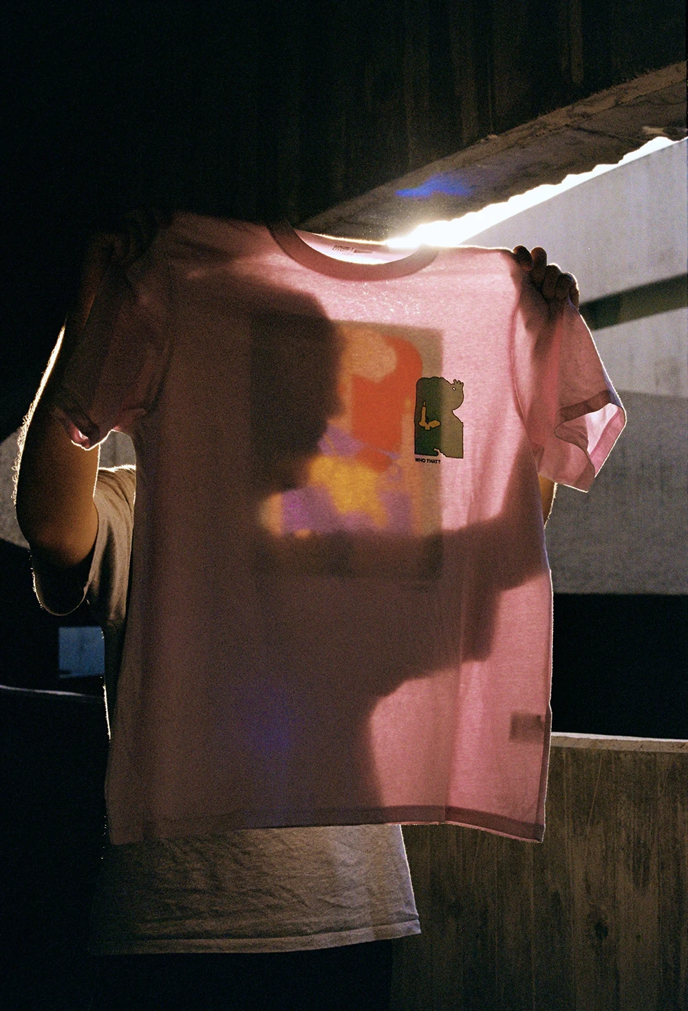A person holding up the Uniqlo x Jhon Boy x Louvre T-shirt against the light, revealing the layered graphic design. The back print, inspired by Vermeer’s “The Lacemaker”, is softly illuminated, emphasizing the interplay of colors and shapes in the reimagined composition. Photograph by Elena Navarro.